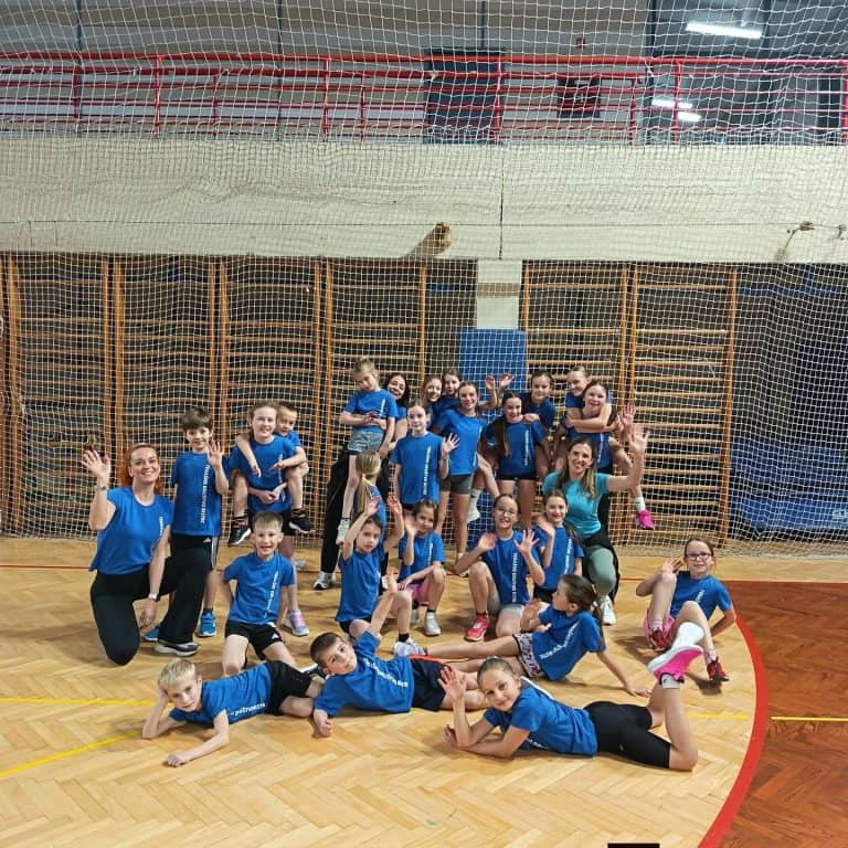 Skupina otrok in trenerjev v športni dvorani, pripravljeni na vadbo.